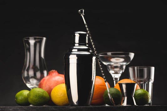 Cocktail shaker with limes, oranges, and glasses on a dark background
